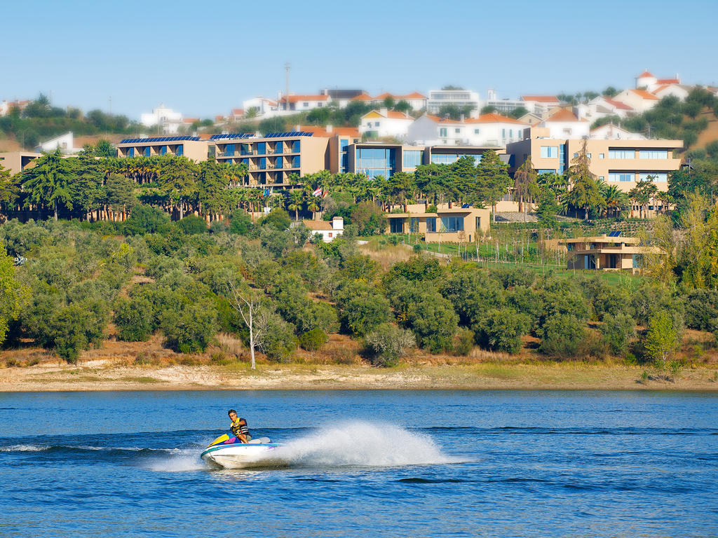 Ap Lago Montargil - Conference & Spa Dış mekan fotoğraf The photo depicts a person riding a jet ski on a body of water. In the background, there are modern buildings and lush greenery, suggesting a scenic landscape, likely near a resort or upscale residential area. The clear blue sky and calm water enhanc