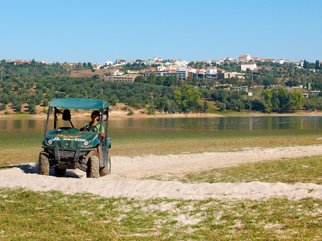 Ap Lago Montargil - Conference & Spa Dış mekan fotoğraf The photo shows a small green all-terrain vehicle driving along a sandy path beside a body of water. In the background, there's a landscape with trees and rolling hills, along with a town or village visible on the hillside. The sky is clear, indicati
