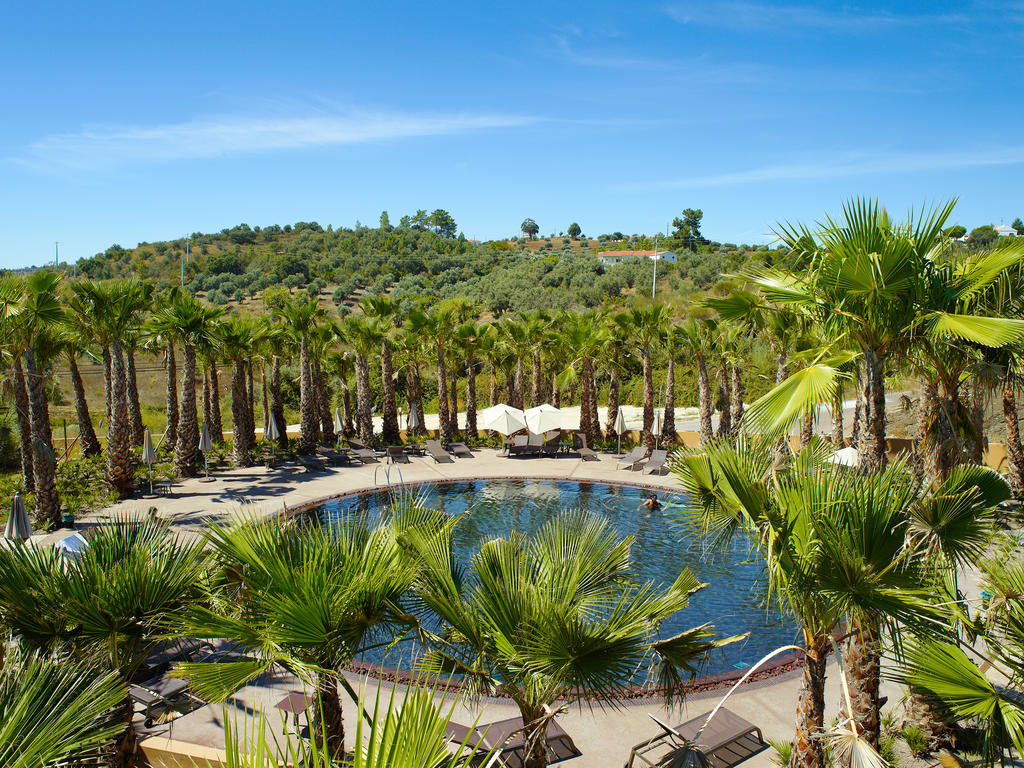 Ap Lago Montargil - Conference & Spa Dış mekan fotoğraf The photo depicts a beautiful outdoor area featuring a circular swimming pool surrounded by palm trees. The pool area has several lounge chairs arranged around it. In the background, there is a lush green hillside, and the sky is clear with a bright 