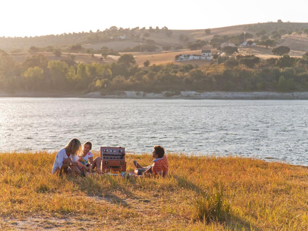 Ap Lago Montargil - Conference & Spa Dış mekan fotoğraf The photo depicts a serene lakeside scene during sunset. Three people are gathered on a grassy area near the water, enjoying a picnic. They are seated around a picnic basket, with the shimmering lake in the background and hills in the distance. The w