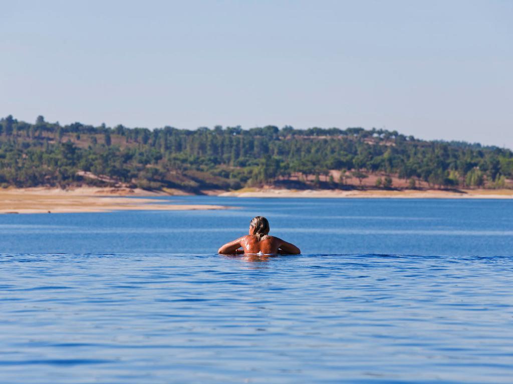 Ap Lago Montargil - Conference & Spa Dış mekan fotoğraf The photo shows a person swimming in a calm body of water, likely a lake or a river. The swimmer is seen from behind and appears to be enjoying the water on a sunny day. In the background, there's a scenic landscape featuring green trees and a sandy 