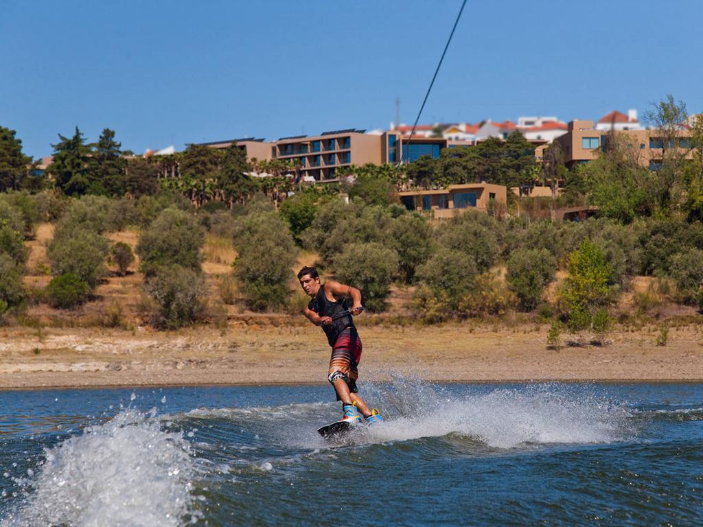 Ap Lago Montargil - Conference & Spa Dış mekan fotoğraf The photo shows a person wakeboarding on a body of water. The individual is being pulled by a cable, which is likely connected to a boat or a winch. The person is wearing a life jacket and swim shorts, balancing on the board as they enjoy the activit