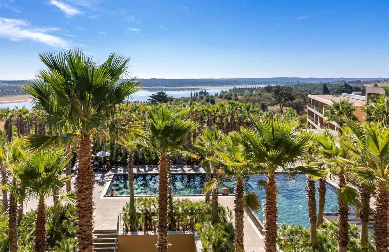 Ap Lago Montargil - Conference & Spa Dış mekan fotoğraf The photo depicts a picturesque resort landscape with a clear blue sky. In the foreground, there are tall palm trees surrounding a swimming pool area. The swimming pool has a modern design and is surrounded by lush greenery. In the background, you ca
