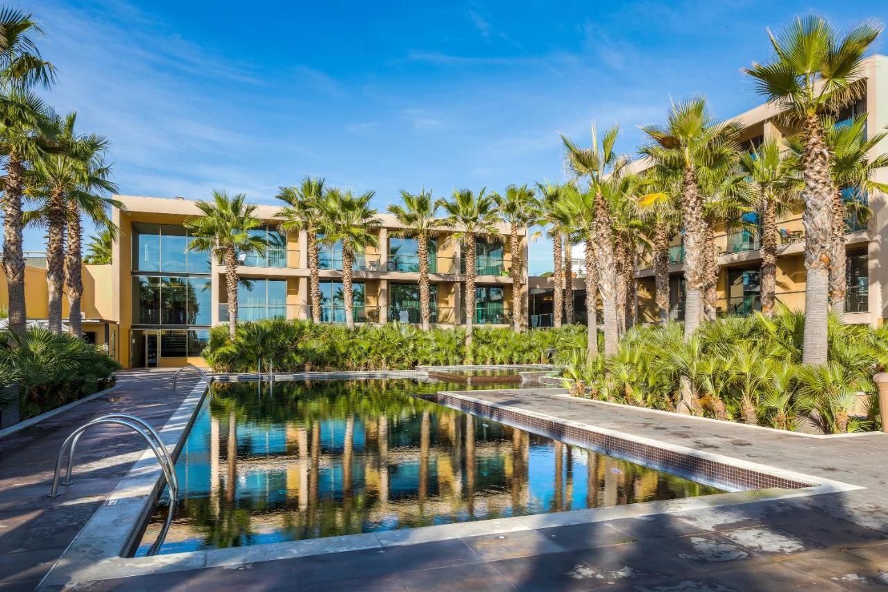 Ap Lago Montargil - Conference & Spa Dış mekan fotoğraf The photo features a modern building surrounded by lush greenery and palm trees. In the foreground, there is a serene swimming pool with a reflective surface that captures the surrounding environment. The architecture of the building appears to have 