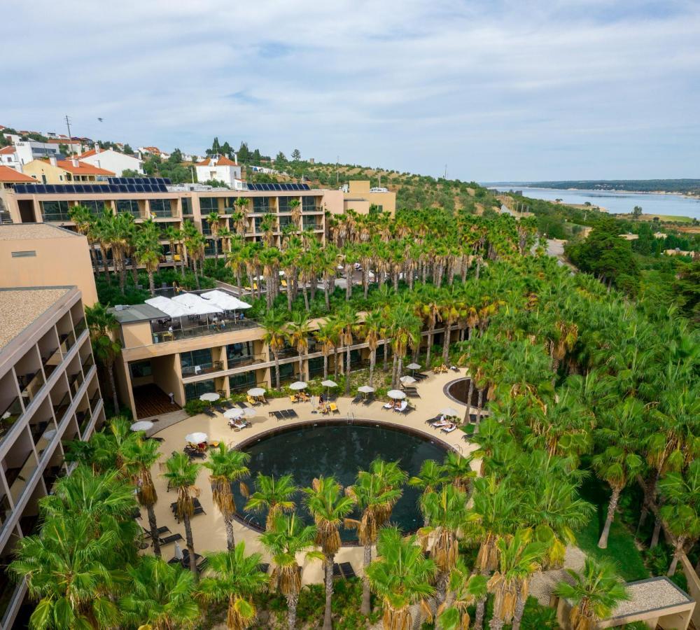 Ap Lago Montargil - Conference & Spa Dış mekan fotoğraf The photo shows a modern hotel complex set amidst lush greenery, featuring numerous palm trees. In the foreground, there is a circular swimming pool surrounded by lounge chairs. The hotel itself has multiple levels with balconies overlooking the pool