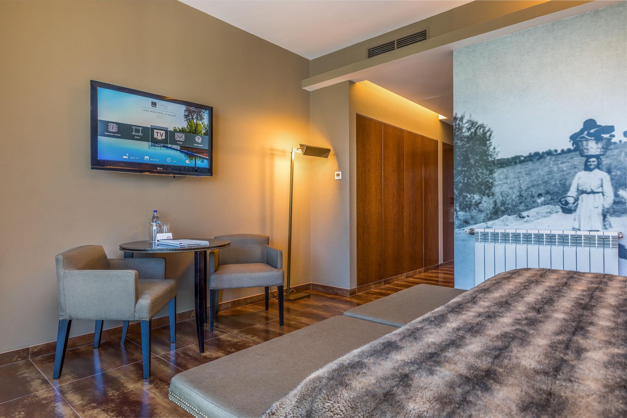 Ap Lago Montargil - Conference & Spa Dış mekan fotoğraf The photo shows an interior of a room, likely a hotel or a modern living space. In the foreground, there is a bed with a textured brown blanket. To one side, there are two comfortable chairs and a small table, possibly with a bottle of water. A flat-