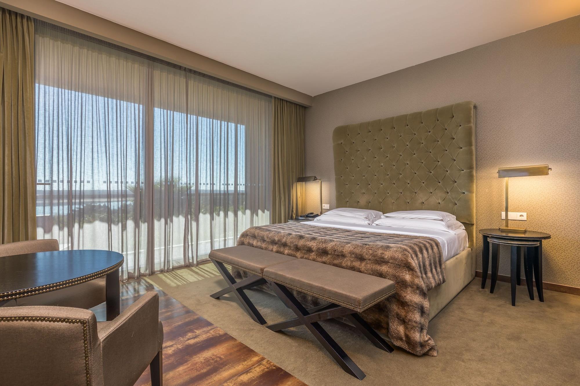 Ap Lago Montargil - Conference & Spa Dış mekan fotoğraf The photo shows a modern hotel room featuring a large bed with a tufted headboard and a cozy blanket. There are curtains draped over large windows that allow natural light to flow in, providing a view of the outside. A small seating area includes two