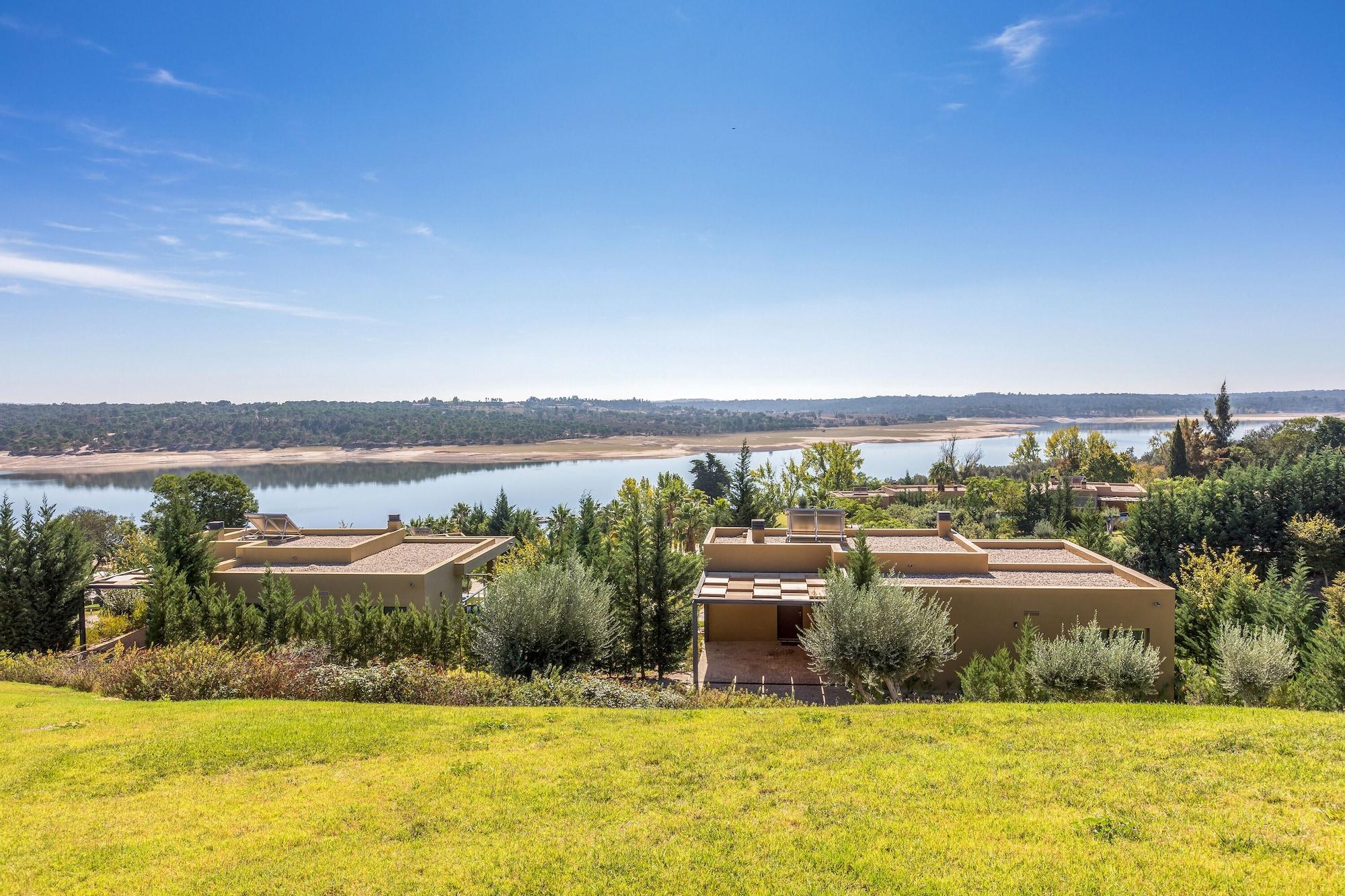 Ap Lago Montargil - Conference & Spa Dış mekan fotoğraf The photo depicts a scenic landscape featuring a calm body of water, possibly a lake or river, surrounded by lush greenery and trees. In the foreground, there are several modern buildings with flat roofs, set against a vibrant blue sky. The overall s