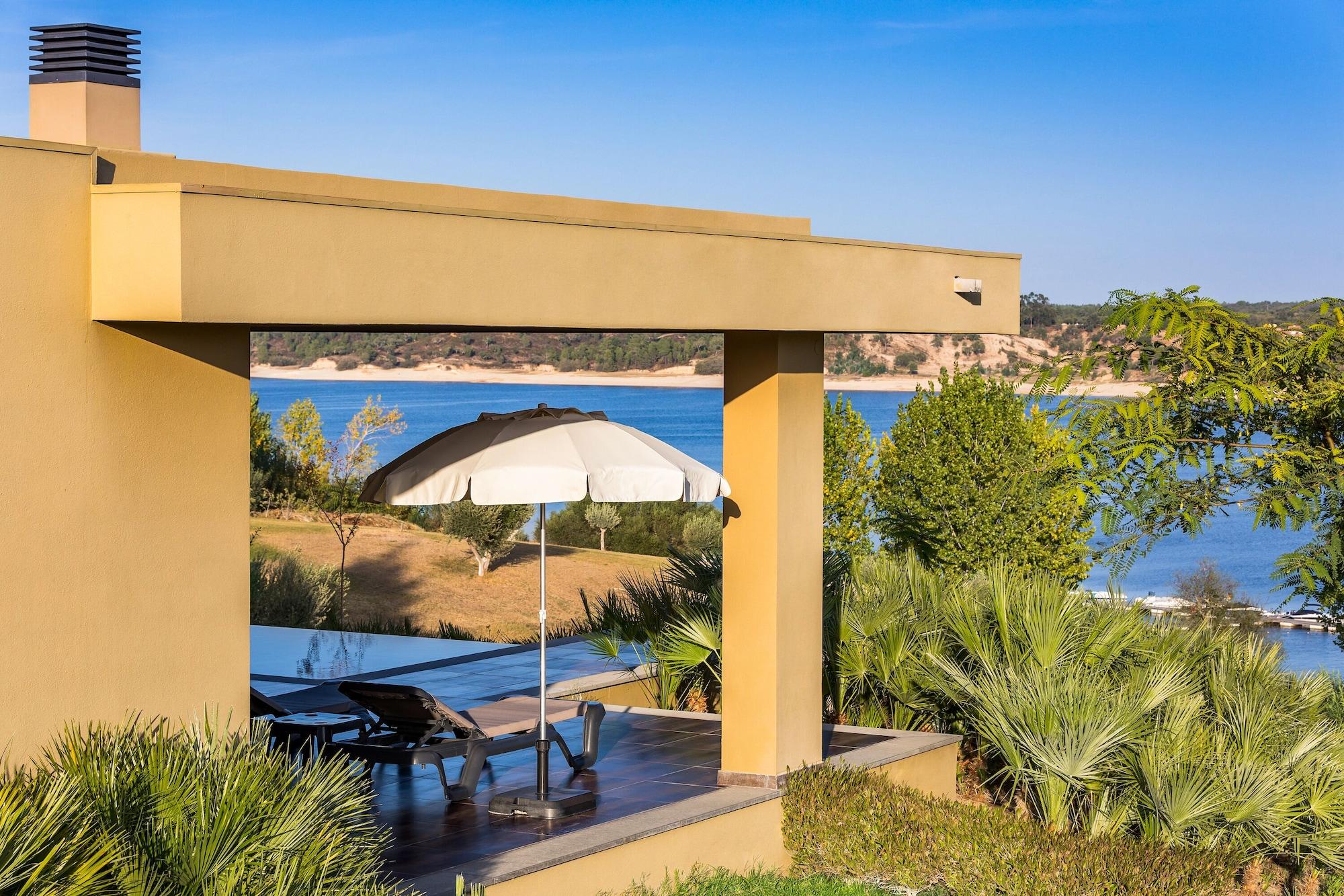 Ap Lago Montargil - Conference & Spa Dış mekan fotoğraf The photo shows a peaceful outdoor area with a modern architectural structure featuring an overhang. There is a lounge chair positioned beneath a large umbrella, creating a shaded spot to relax. In the background, there is a picturesque view of a bod