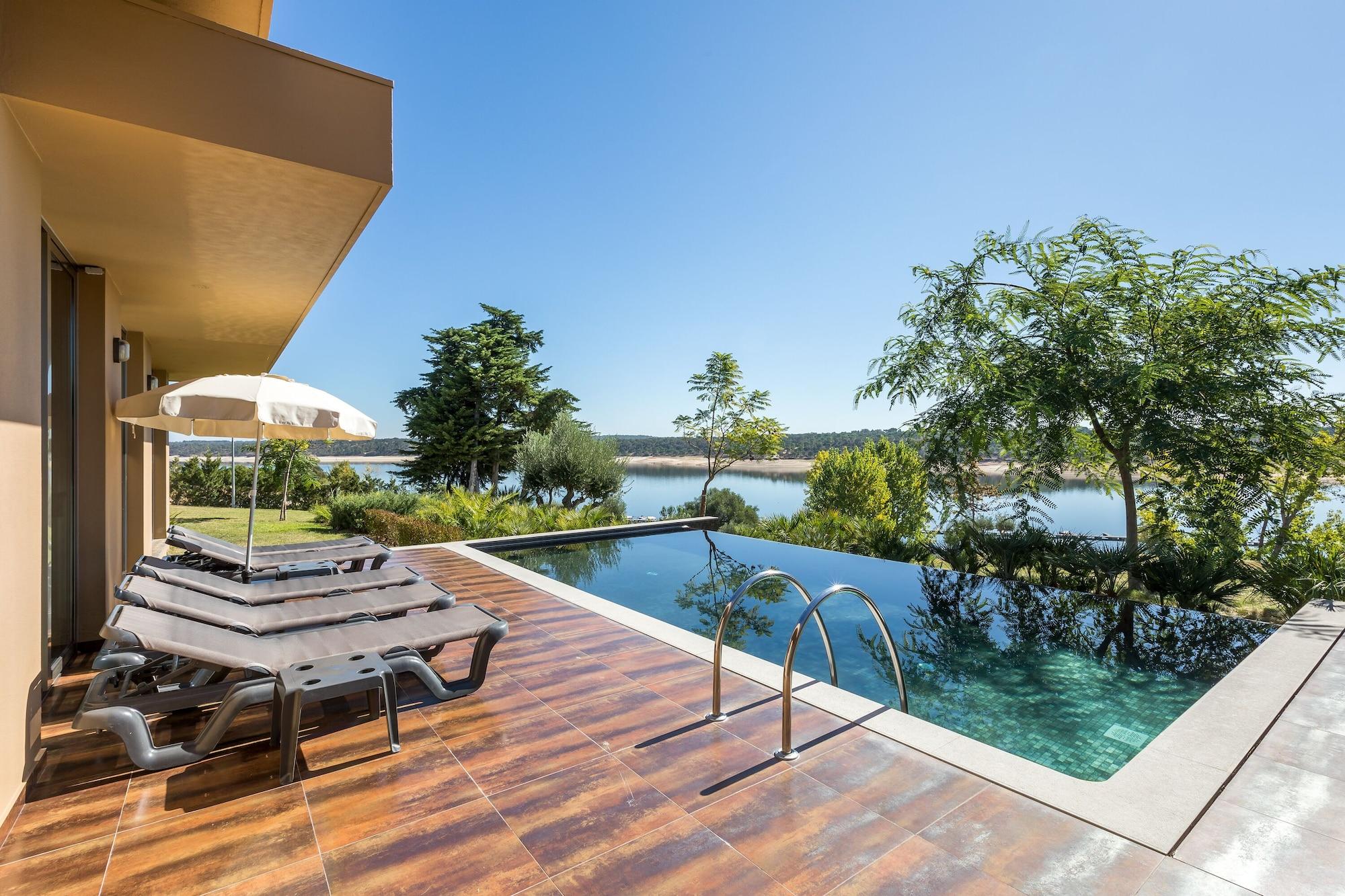 Ap Lago Montargil - Conference & Spa Dış mekan fotoğraf The photo shows a luxurious outdoor setting by a pool. There are several lounge chairs arranged next to an infinity pool, which overlooks a scenic body of water and lush greenery. A parasol provides shade for relaxation. The sky is clear and blue, in