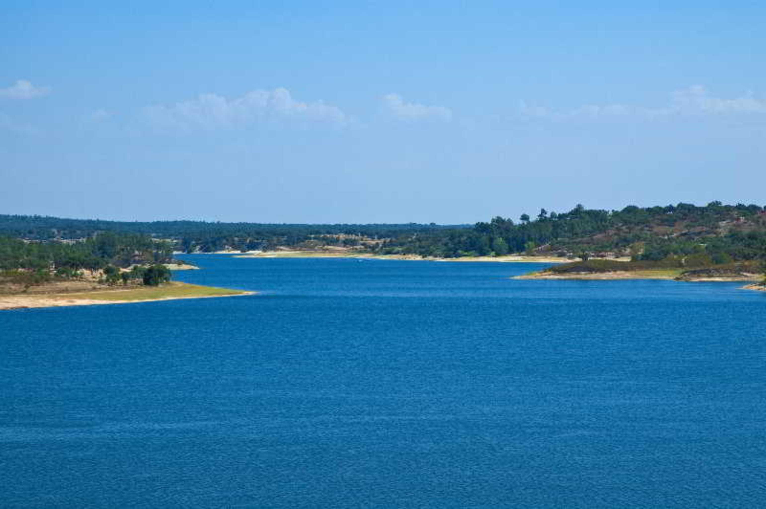 Ap Lago Montargil - Conference & Spa Dış mekan fotoğraf The photo depicts a serene landscape featuring a large body of water, likely a lake or river. The water appears calm and is reflecting a clear blue sky with some clouds. Surrounding the water, there are patches of green land and trees, indicating a n