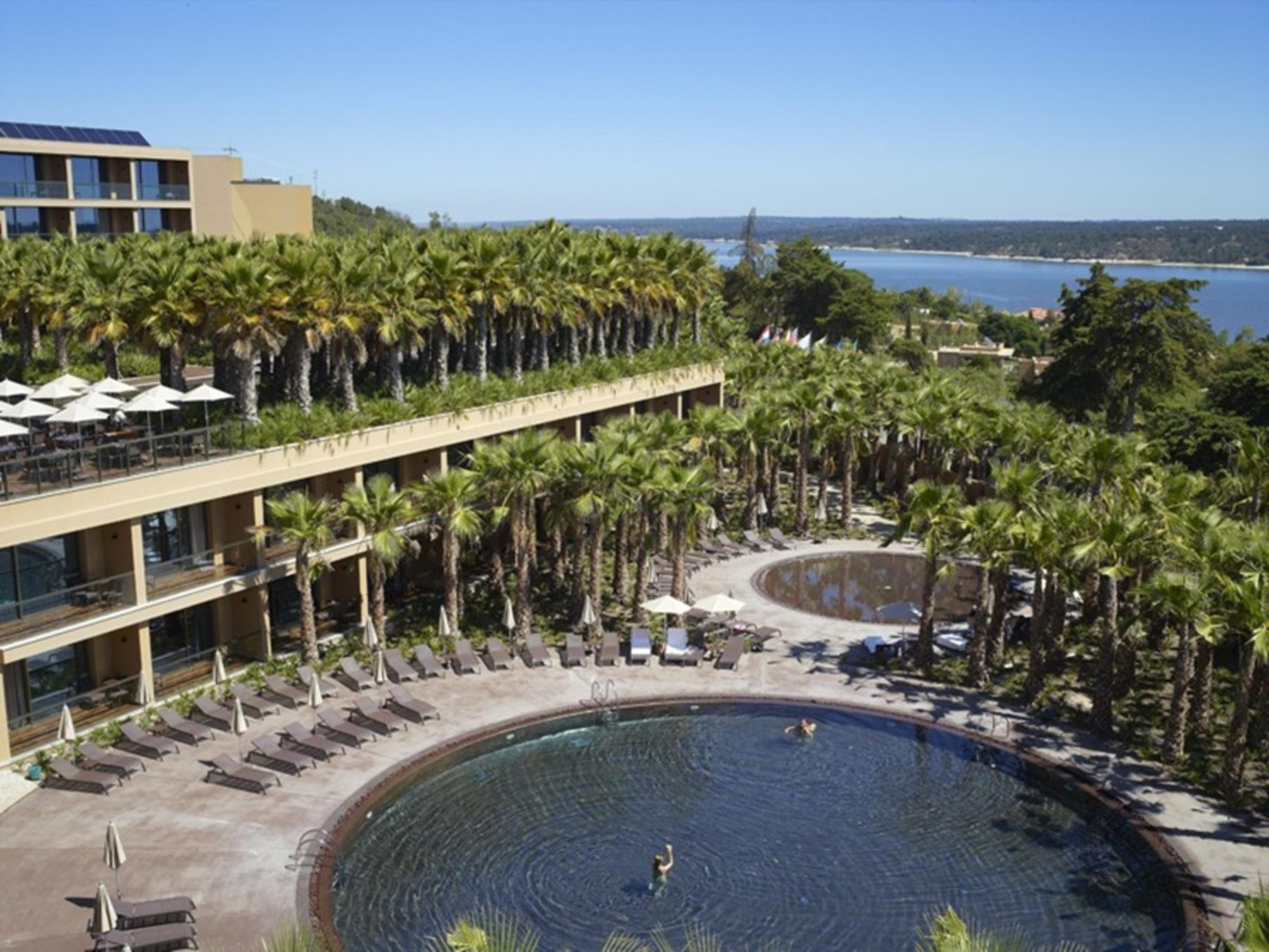 Ap Lago Montargil - Conference & Spa Dış mekan fotoğraf The photo shows a scenic view of a resort area. In the foreground, there are circular pools surrounded by lounge chairs, with a few people enjoying the water. Palm trees line the edges, creating a tropical atmosphere. In the background, there is a mu