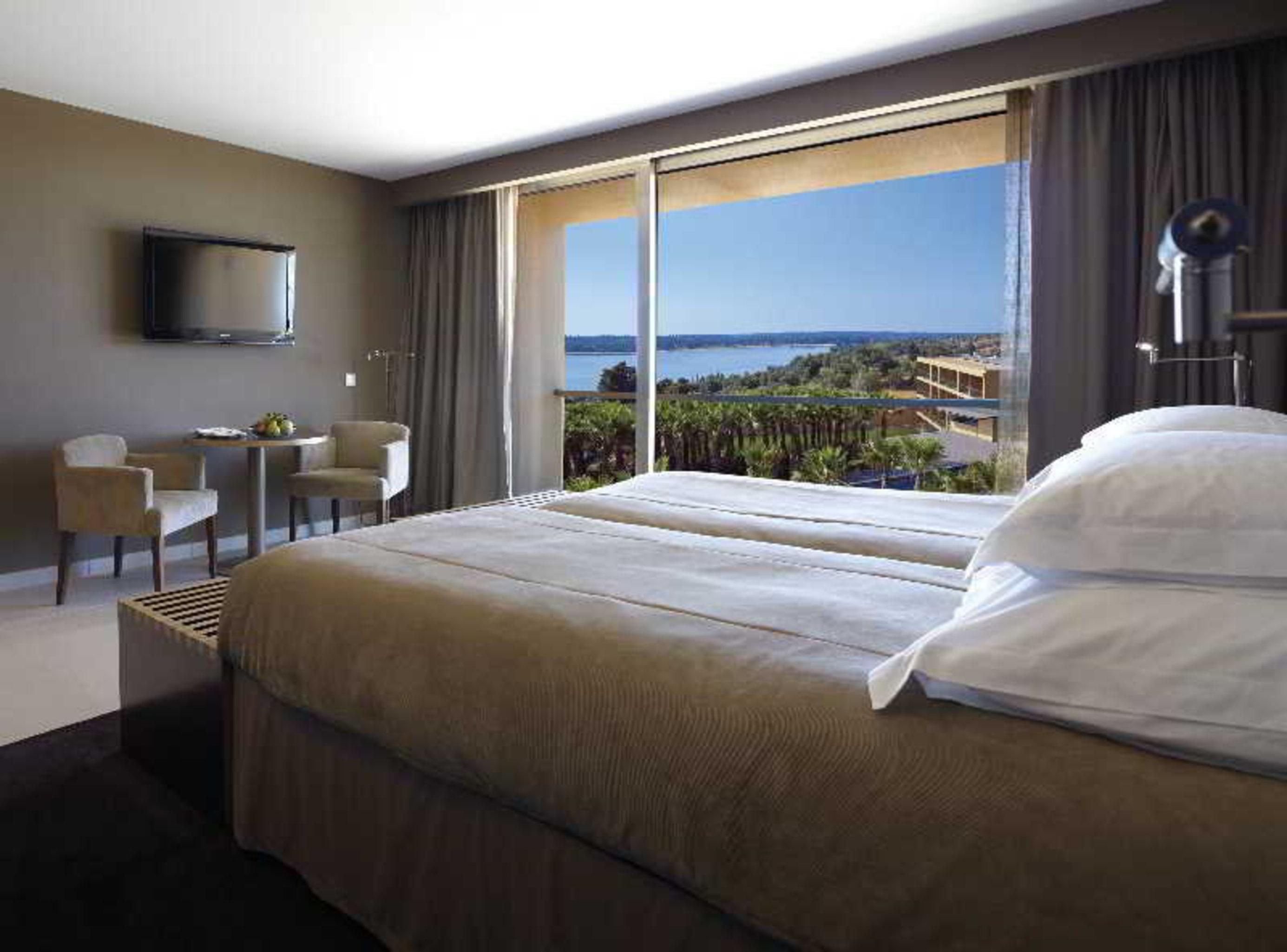 Ap Lago Montargil - Conference & Spa Dış mekan fotoğraf The photo shows a modern hotel room with a large window offering a view of the sea and mountains. The room features a comfortable bed with white pillows and brown bedding. There is a small sitting area with two chairs and a table, as well as a wall-m