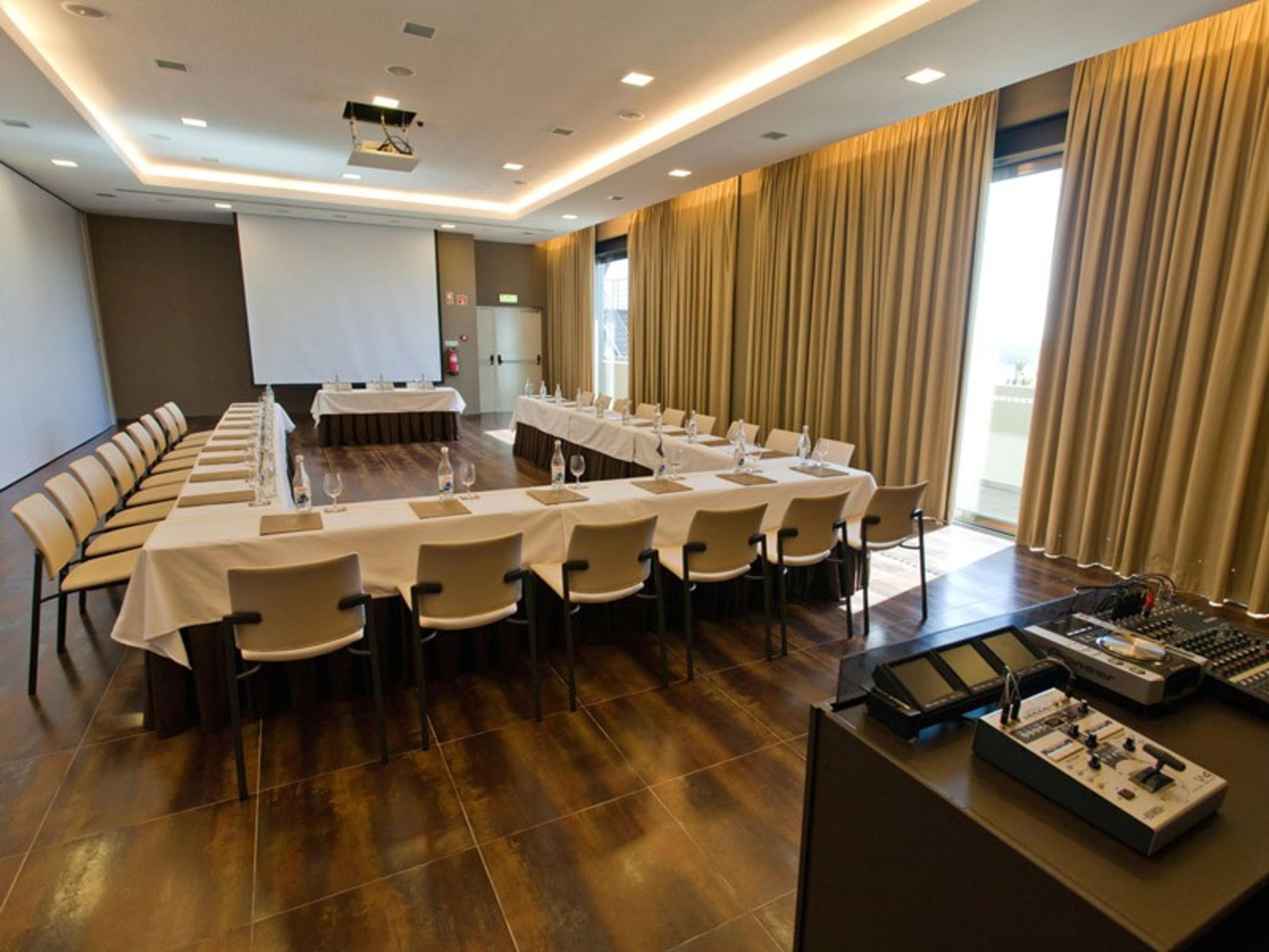 Ap Lago Montargil - Conference & Spa Dış mekan fotoğraf The photo shows a modern conference room set up for a meeting. There are several tables arranged in a U-shape, surrounded by chairs. A large screen and projector are positioned at the front, with a table for speakers or presenters. The room has large