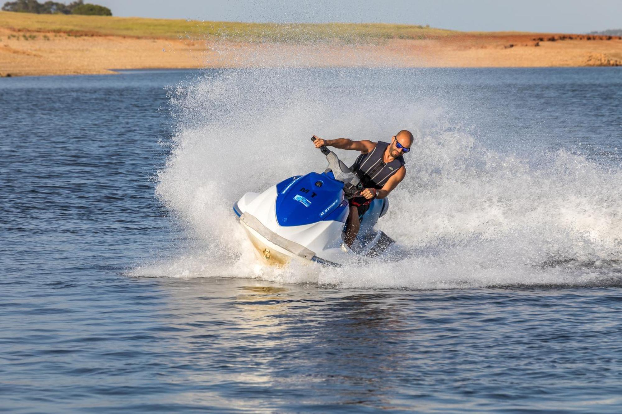 Ap Lago Montargil - Conference & Spa Dış mekan fotoğraf The photo shows a person riding a jet ski over a body of water. The rider is wearing sunglasses and a life vest, leaning forward as the jet ski accelerates, creating a spray of water around them. The background features a natural landscape, likely a 