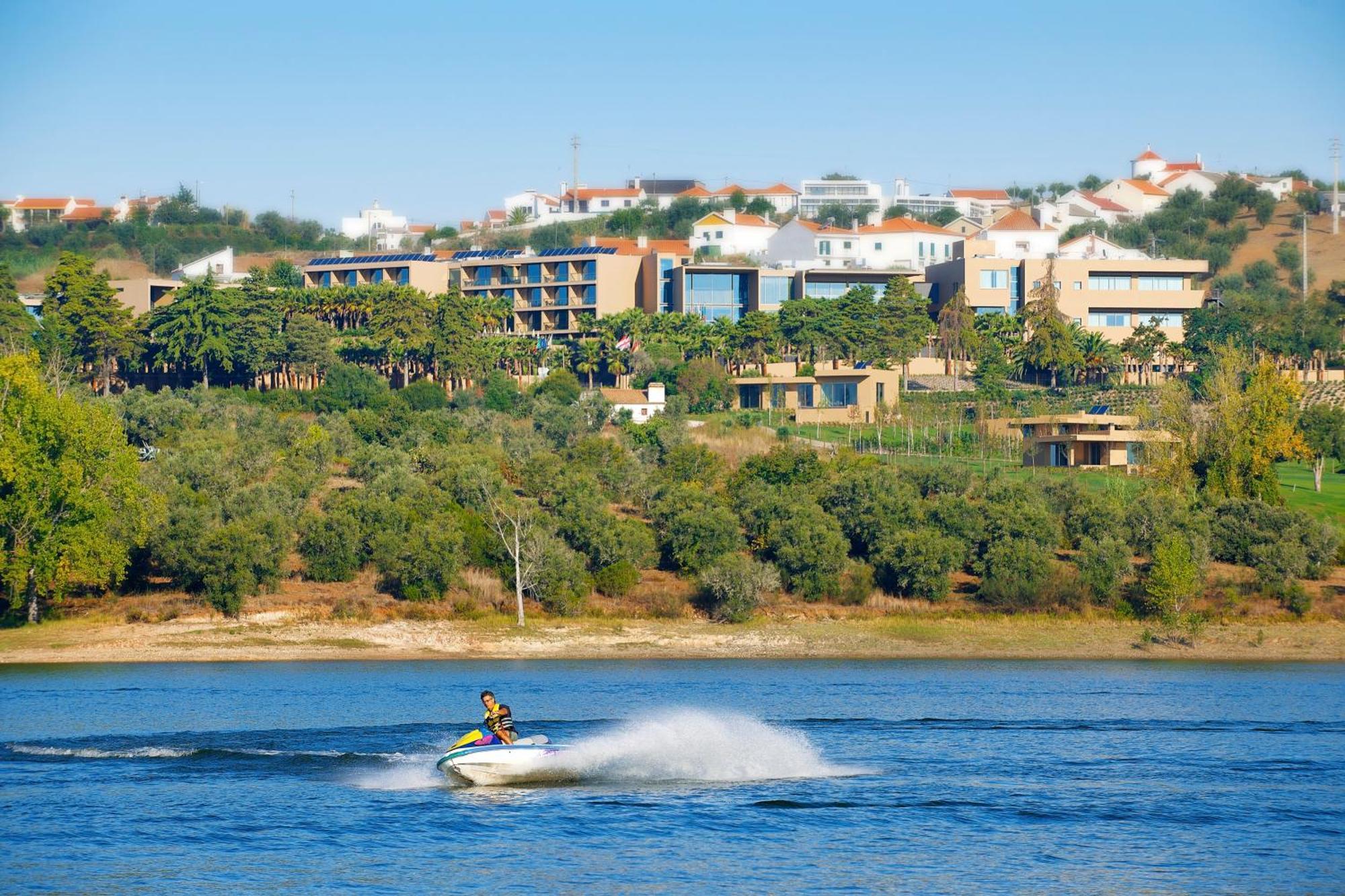 Ap Lago Montargil - Conference & Spa Dış mekan fotoğraf The photo shows a person riding a jet ski on a body of water, creating a spray as they speed across the surface. In the background, there are modern buildings and a hillside with trees and greenery. The sky is clear and blue, suggesting a bright and 