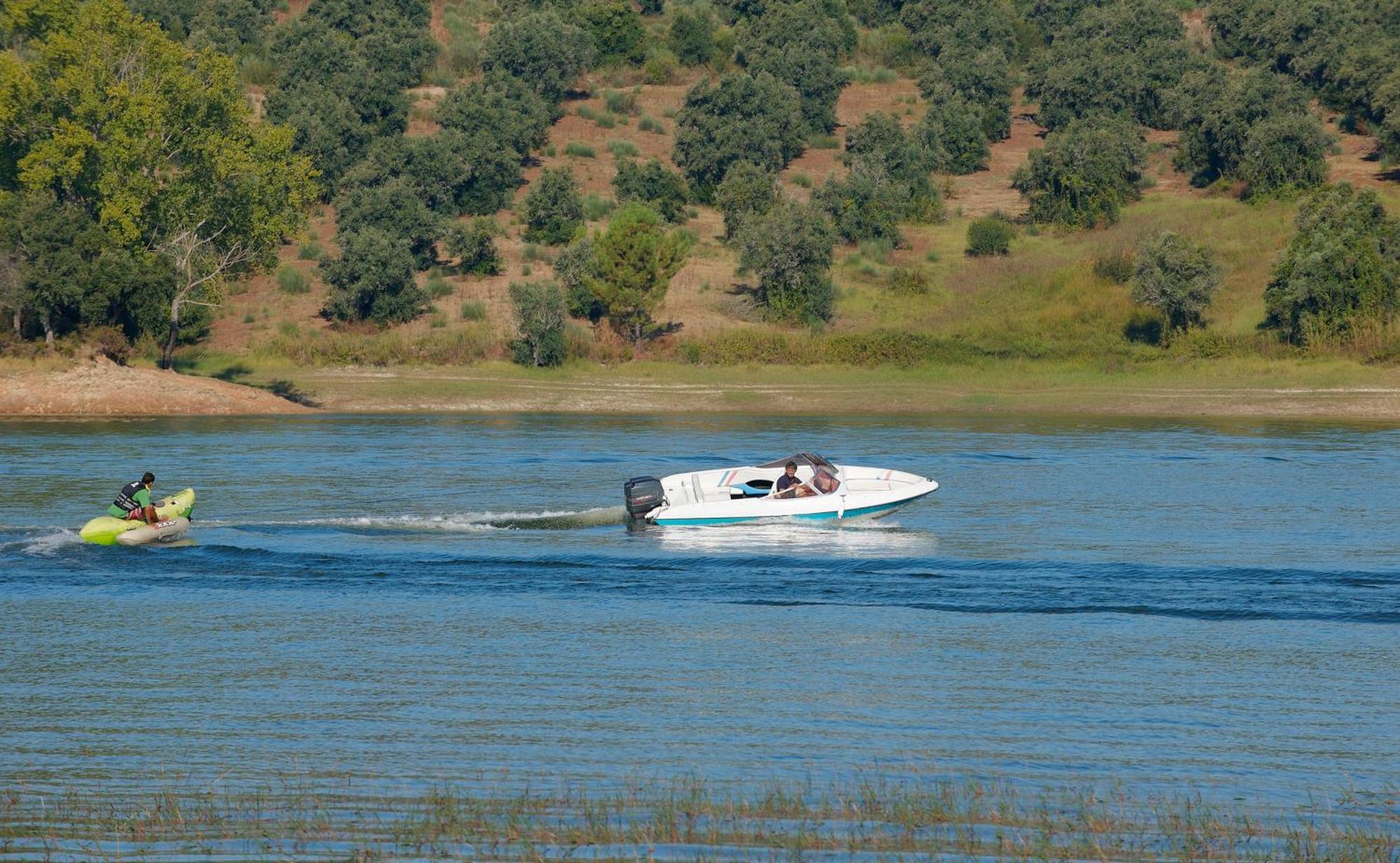Ap Lago Montargil - Conference & Spa Dış mekan fotoğraf The photo shows a scene on a water body, likely a river or lake. In the foreground, a speedboat is moving quickly through the water, leaving a wake behind it. There are two people visible in the boat. In the background, there is a hilly area covered 