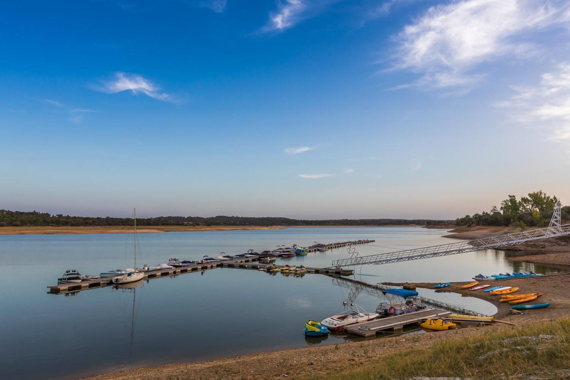 Ap Lago Montargil - Conference & Spa Dış mekan fotoğraf The photo shows a serene lakeside scene. In the foreground, there are several boats moored at a dock, with some colorful kayaks and canoes lined up along the shore. The water is calm and reflects the sky, which features a few wispy clouds. In the bac