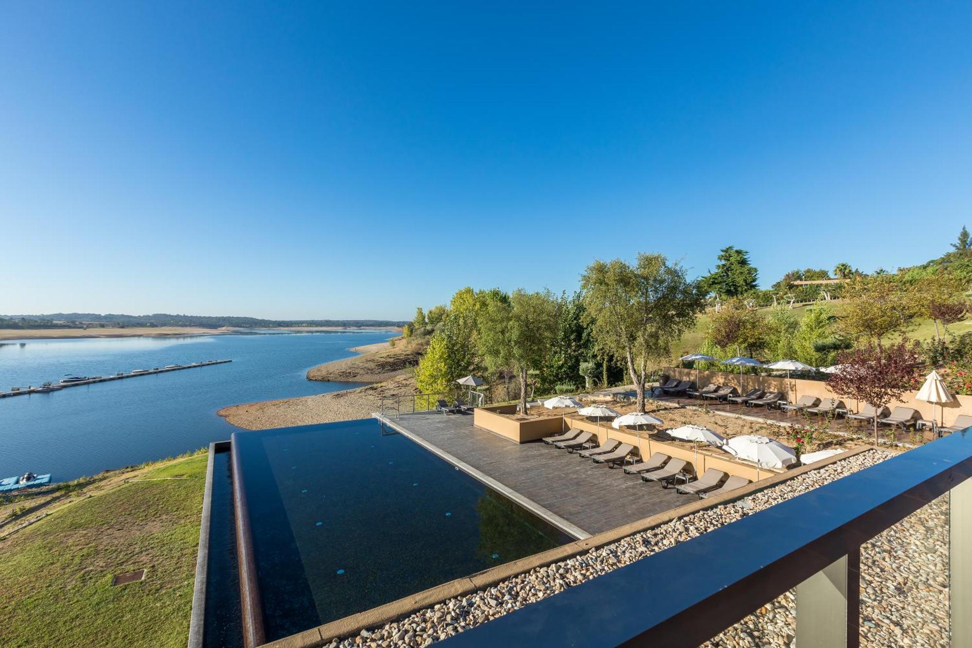 Ap Lago Montargil - Conference & Spa Dış mekan fotoğraf The photograph depicts a scenic view of a tranquil water body, likely a lake or river, bordered by lush greenery. In the foreground, there is an infinity pool that blends seamlessly with the water beyond. Comfortable lounge chairs are arranged on a w