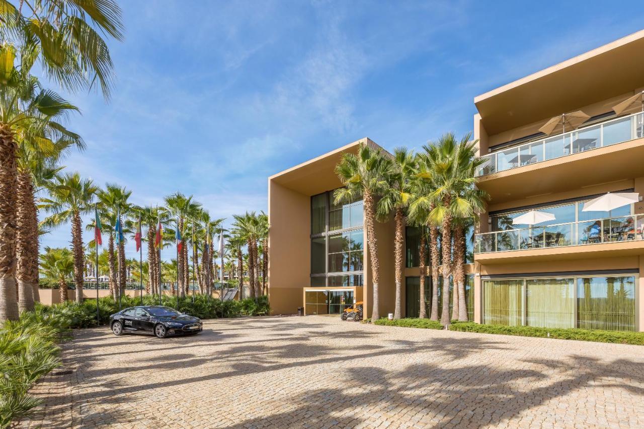 Ap Lago Montargil - Conference & Spa Dış mekan fotoğraf The photo shows a modern building with a sleek design, featuring large glass windows and balconies. The structure is surrounded by a well-maintained outdoor area with palm trees, adding a tropical feel. In front of the building, there is a paved park